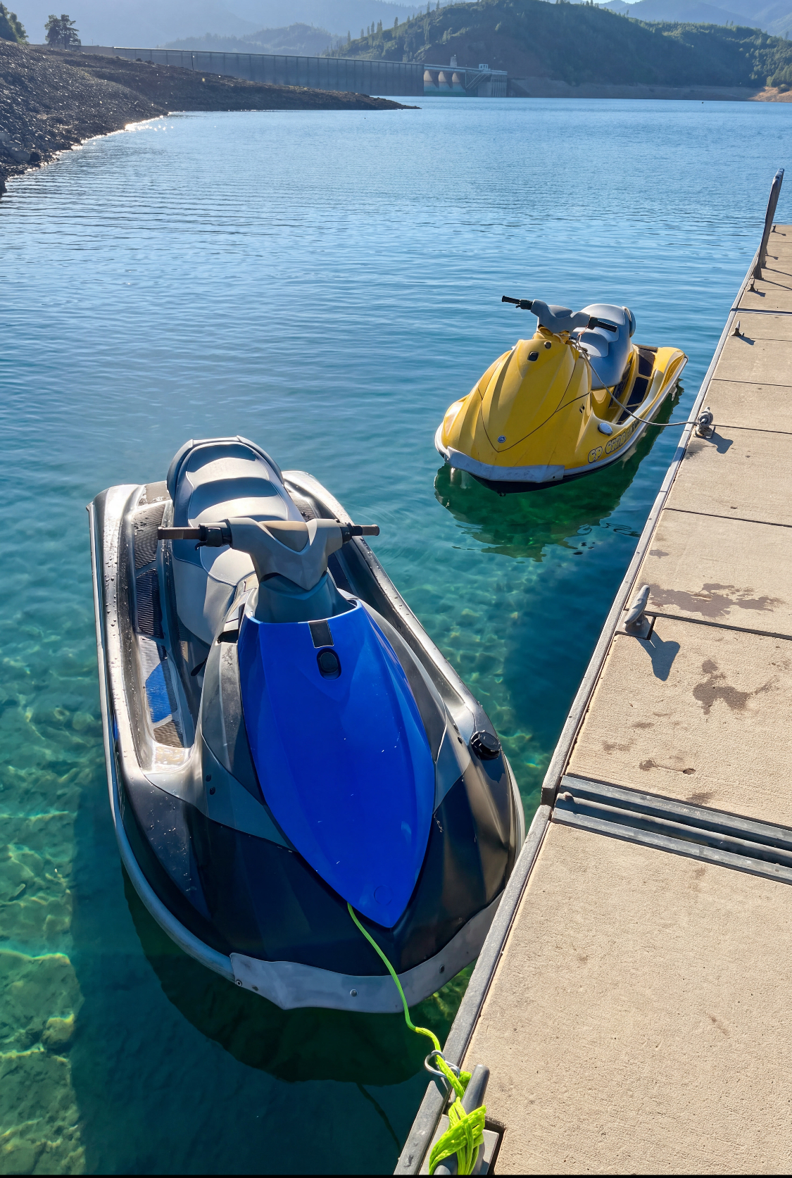 The blue and the yellow WaveRunner tied along the dock on glassy, crystal-clear turquoise water with Shasta Dam behind