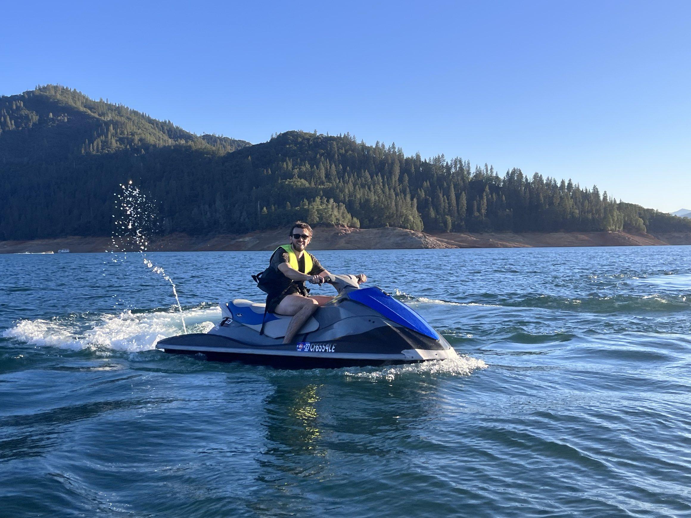 A smiling rider in sunglasses and a yellow life vest cruising the blue WaveRunner with a rooster tail of spray and forested hills behind