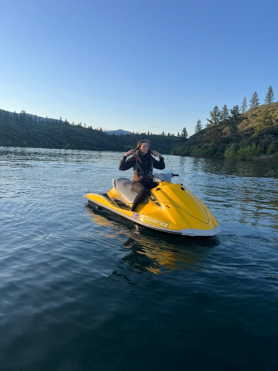 A rider sitting on the yellow jet ski on perfectly glassy water with a pine ridgeline reflected behind