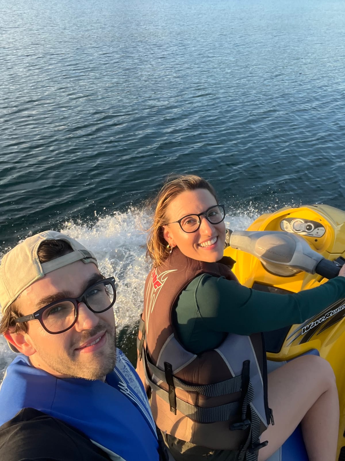 Two riders in life jackets smiling back over their shoulders while riding the yellow WaveRunner