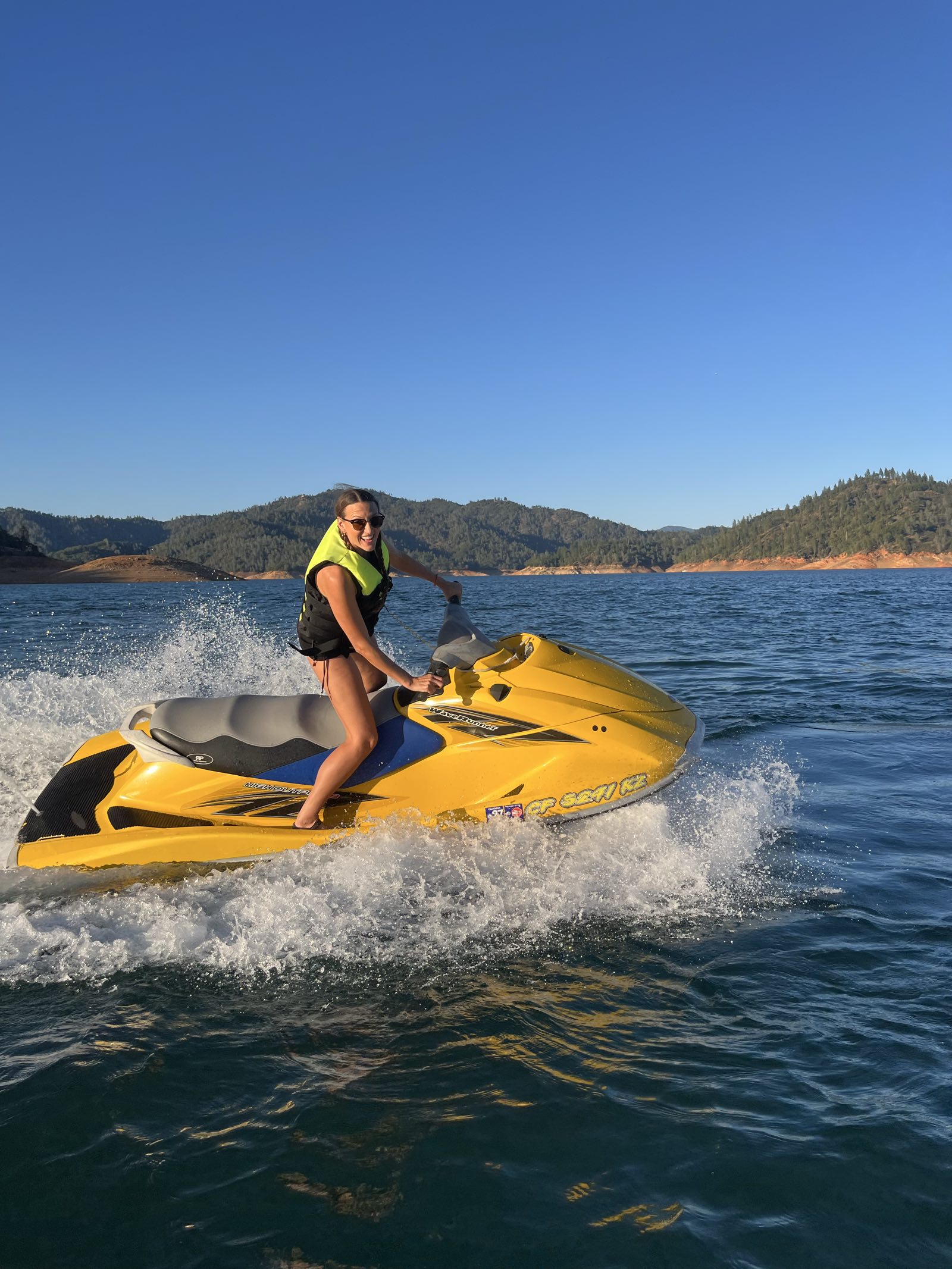 A rider in a yellow life vest leaning into a fast turn on the yellow WaveRunner with big spray, mountains and blue sky behind