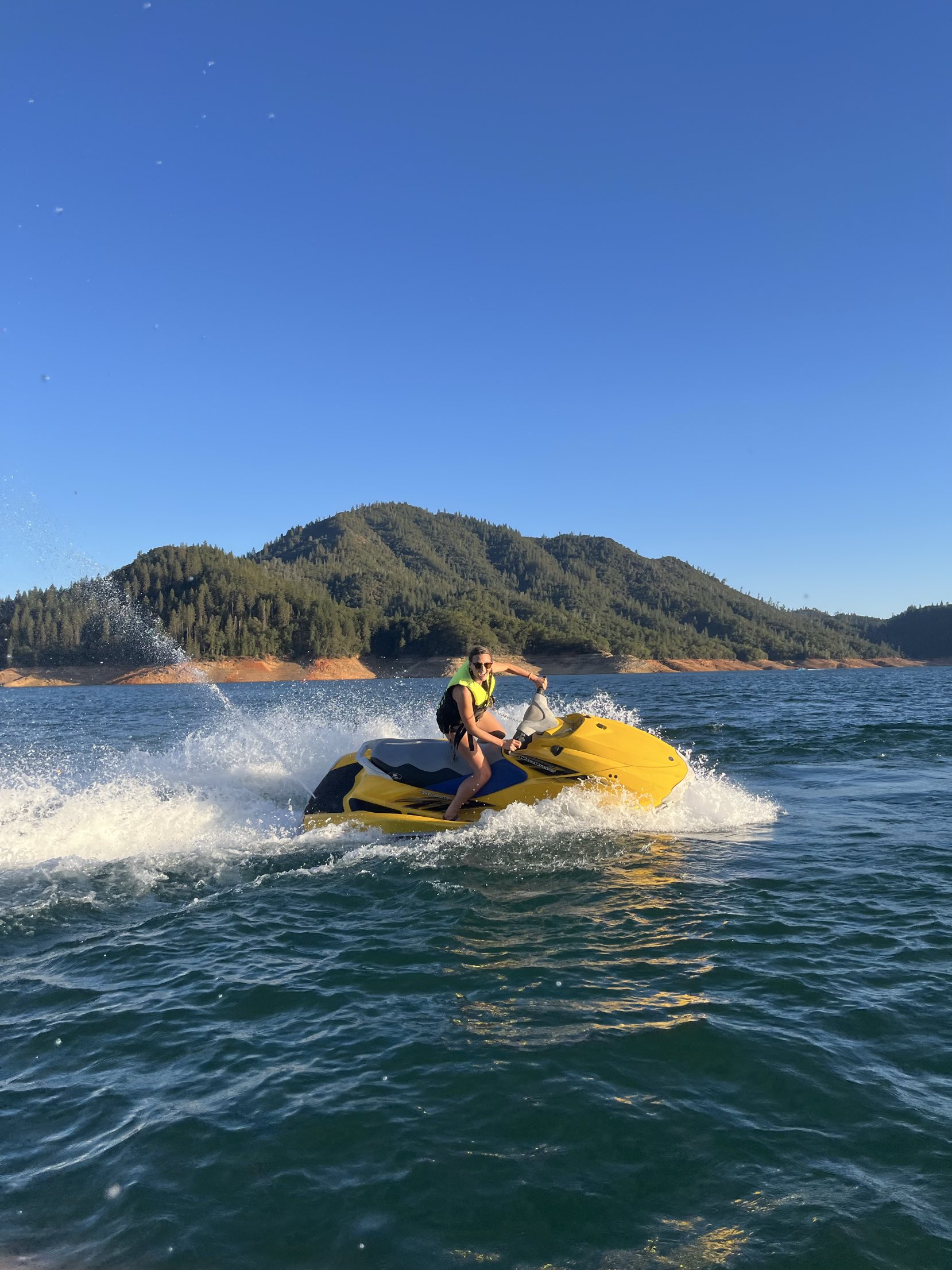 A rider in a yellow life vest carving the yellow WaveRunner through a spray-filled turn with forested Shasta hills behind